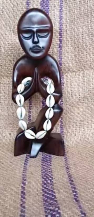 Small dark wooden kneeling figure with hands together holding a cowrie bracelet
